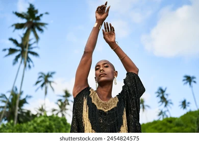 Androgenous black fashion model stands on scenic tropical palm beach portrait. Non-binary ethnic person wearing makeup, jewellery poses with hands up at exotic location. Trans sexual. Pride Stock Photo